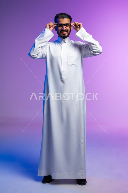 الاناقة والاهتمام بالمظهر الخارجي، بورتريه لرجل عربي خليجي سعودي مبتسم يرتدي الثوب التقليدي يمسك بيده نظاره طبيه ينظر إلى الكاميرا بإيماءات السرور والسعادة، ارتداء النظارات الخاصة بضعف النظر، مفهوم تحسين الرؤية ومحاولة تعديل البصر، صورة لكامل الجسم، خلفية ملونة
