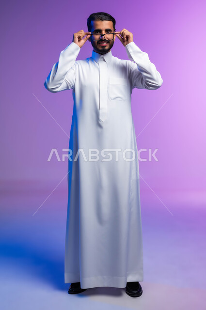 الاناقة والاهتمام بالمظهر الخارجي، بورتريه لرجل عربي خليجي سعودي مبتسم يرتدي الثوب التقليدي يمسك بيده نظاره طبيه ينظر إلى الكاميرا بإيماءات السرور والسعادة، ارتداء النظارات الخاصة بضعف النظر، مفهوم تحسين الرؤية ومحاولة تعديل البصر، صورة لكامل الجسم، خلفية ملونة