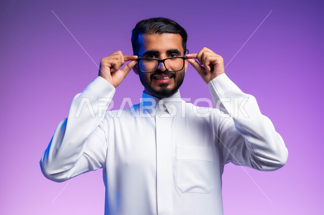Looking at the camera with gestures of pleasure and happiness, a portrait of a Saudi Gulf Arab man wearing a traditional dress, holding glasses in his hand, taking care of the external appearance, wearing glasses for low vision, the concept of improving vision and trying to correct eyesight, a colored background.