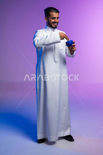 الاهتمام بالمظهر الخارجي، بورتريه لرجل عربي خليجي سعودي يرتدي الثوب التقليدي يمسك بيده كريم الترطيب، مفهوم العناية بنضارة وتحسين البشرة، استخدام كريمات ومكونات تجميلية، صورة الجسم بالكامل، خلفية ملونة