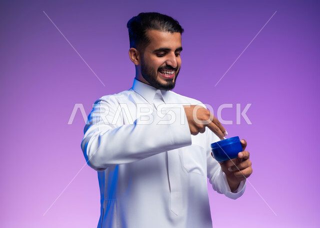 مفهوم العناية بنضارة وتحسين البشرة، الاهتمام بالمظهر الخارجي، بورتريه لرجل عربي خليجي سعودي يرتدي الثوب التقليدي يمسك بيده كريم الترطيب بإيماءات السرور، استخدام كريمات ومكونات تجميلية، صورة الجسم بالكامل، خلفية ملونة