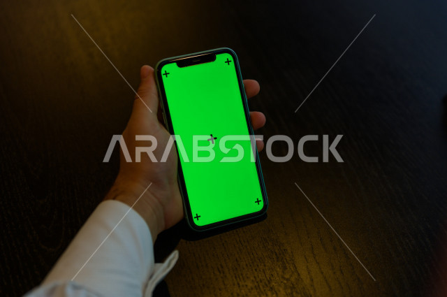 رجل أعمال سعودي خليجي يستخدام هاتفه المحمول بخلفية خضراء كروما ، تقنية و حاسب ، استخدام تقنية السمارت على الهاتف المحمول ليتحكم بالتلفاز