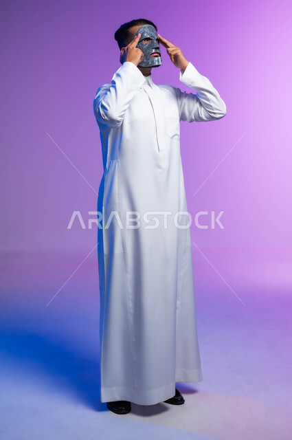 استخدام مكونات تجميلية، خطوات تنظيف وتقشير البشرة، بورتريه لرجل عربي خليجي سعودي يقوم بوضع قناع الطين على وجهه، مفهوم العناية والاهتمام بالمظهر الخارجي، صورة الجسم بالكامل، خلفية بيضاء.