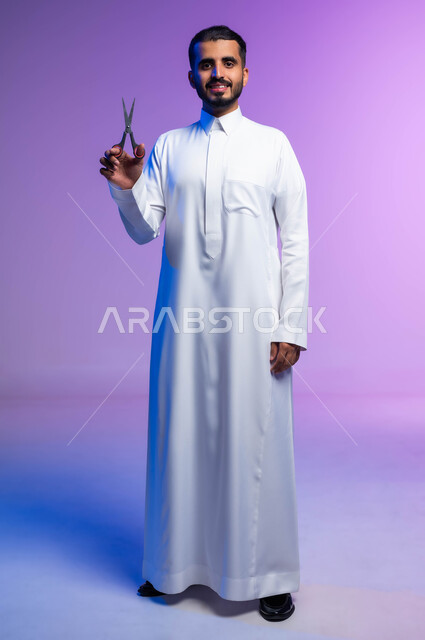 النظر إلى الكاميرا بإيماءات السرور، مركز تجميلي رجالي، مفهوم العناية و الاهتمام بالجمال والمظهر الخارجي، قص و تصفيف الذقن والشارب في صالون الحلاقة، بورتريه لرجل عربي خليجي يرتدي الثوب السعودي يحمل بيده المقص، صورة لكامل الجسم، خلفية ملونة.