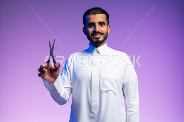 النظر إلى الكاميرا بإيماءات السرور، مركز تجميلي رجالي، مفهوم العناية و الاهتمام بالجمال والمظهر الخارجي، قص و تصفيف الذقن والشارب في صالون الحلاقة، بورتريه مقرب لرجل عربي خليجي يرتدي الثوب السعودي يحمل بيده المقص، خلفية ملونة.