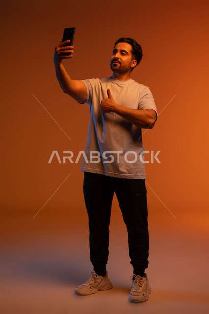 إجراء مكالمات الفيديو, الاعجاب بآداء التقنية الحديثة، التقاط صور السيلفي التذكارية، بورتريه مقرب لشاب عربي سعودي خليجي مبتسم يرتدي الزي الكاجوال يمسك بيده الهاتف المحمول يرفع إصبع الابهام نحو الأعلى بعلامة الجودة، صورة الجسم بالطول الكامل، خلفية ملونة.