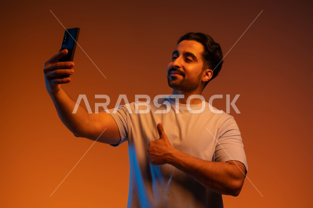 التقاط صور السيلفي التذكارية، الاعجاب بآداء التقنية الحديثة، الشعور بالموافقه والقبول، بورتريه مقرب لشاب عربي سعودي خليجي مبتسم يرتدي الزي الكاجوال يمسك بيده الهاتف المحمول يرفع إصبع الابهام نحو الأعلى بعلامة الجودة، إجراء مكالمات الفيديو, خلفية ملونة.