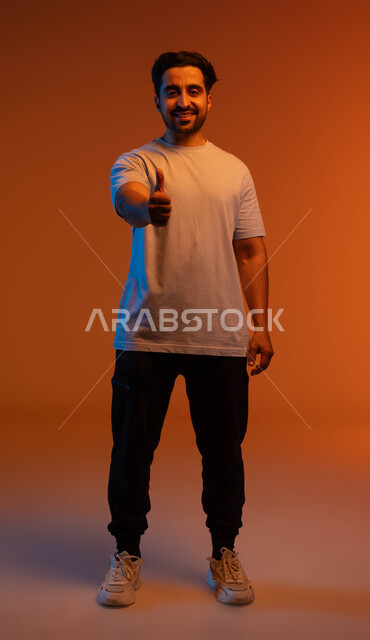 الشعور بالموافقه والقبول، إيماءات الإعجاب والرضى, بورتريه  لشاب عربي سعودي خليجي مبتسم ينظر إلى الكاميرا يرتدي الزي الكاجوال يرفع إصبع الابهام نحو الأعلى بعلامة الجودة، تعابير الإمتياز والتميز, صورة الجسم بالطول الكامل، خلفية ملونة.