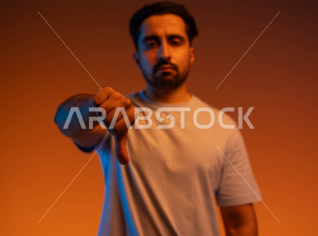 الشعور بعدم القبول والرضا، تعابير الرفض والتقييم السلبي، القلق والفشل في أمر ما, بورتريه مقرب  لرجل عربي خليجي سعودي ينظر إلى الكاميرا يرتدي الزي الكاجوال يرفع إبهامه ويشير للأسفل بإيماءات الإنزعاج, خلفية ملونة.
