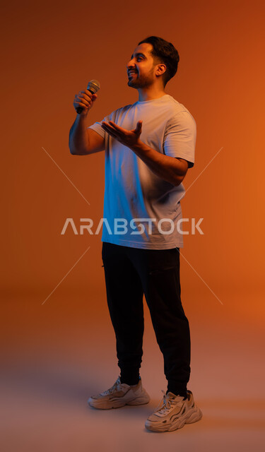 الاستمتاع بأداء المواهب الصوتية، بورتريه مقرب لشاب عربي خليجي سعودي يرتدي الزي الكاجوال يمسك بيده المايكروفون و يغني بحماس، ايماءات تدل على الاندماج والتفاعل مع الغناء، ممارسة موهبة الغناء، تسجيل مقطع صوتي غنائي، صورة الجسم بالطول الكامل، خلفية ملونة.