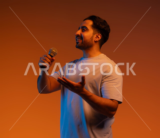 الاستمتاع بأداء المواهب الصوتية، بورتريه مقرب لشاب عربي خليجي سعودي يرتدي الزي الكاجوال يمسك بيده المايكروفون و يغني بحماس، ايماءات تدل على الاندماج والتفاعل مع الغناء، ممارسة موهبة الغناء، تسجيل مقطع صوتي غنائي، خلفية ملونة