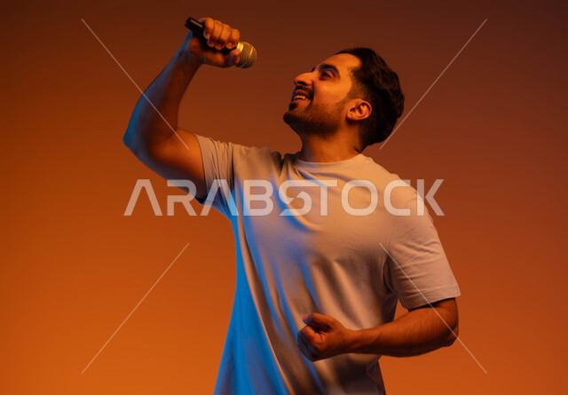 الاستمتاع بممارسة المواهب الفنية، ايماءات تدل على الاندماج والتفاعل مع الغناء، تسجيل مقطع صوتي غنائي، قضاء وقت ممتع  بأداء العروض الصوتية، بورتريه مقرب  لشاب عربي خليجي سعودي يرتدي الزي الكاجوال يمسك بيده المايكروفون و يغني بحماس، خلفية ملونة