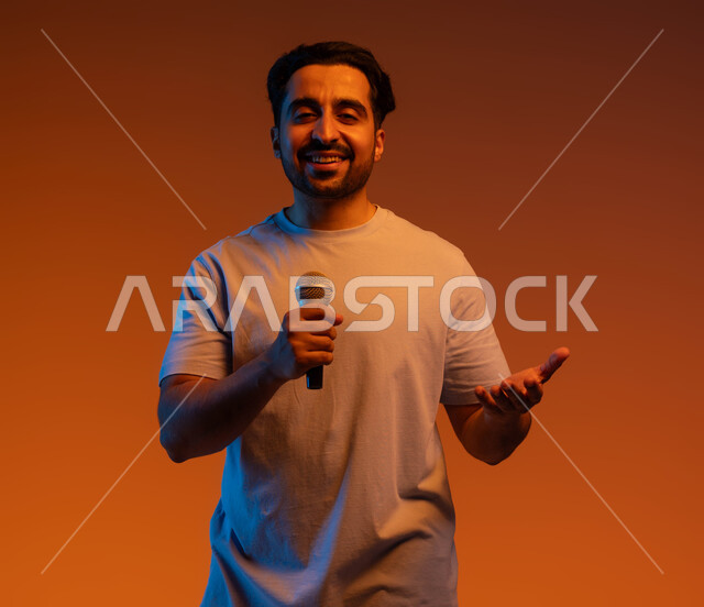 قضاء وقت ممتع  بأداء العروض الصوتية، بورتريه مقرب لشاب عربي خليجي سعودي يرتدي الزي الكاجوال يمسك بيده المايكروفون و يغني بحماس، ايماءات تدل على الاندماج والتفاعل مع الغناء، ممارسة موهبة الغناء، تسجيل مقطع صوتي غنائي، خلفية ملونة