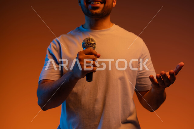 تسجيل مقطع صوتي غنائي، قضاء وقت ممتع  بأداء العروض الصوتية، بورتريه مقرب لشاب عربي خليجي سعودي يرتدي الزي الكاجوال يمسك بيده المايكروفون و يغني بحماس، ايماءات تدل على الاندماج والتفاعل مع الغناء، ممارسة موهبة الغناء، خلفية ملونة