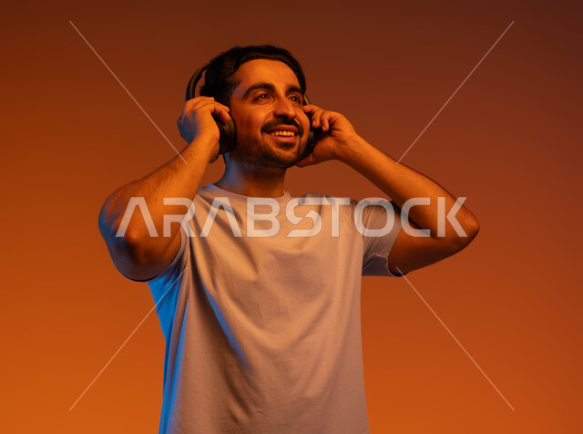 Spending enjoyable times listening to songs and audio clips, a close-up portrait of a smiling Saudi Gulf Arab young man wearing a casual outfit and wearing an audio headset, the concept of listening and hearing using headphone technology, expressions and gestures of pleasure and joy, a colorful background.