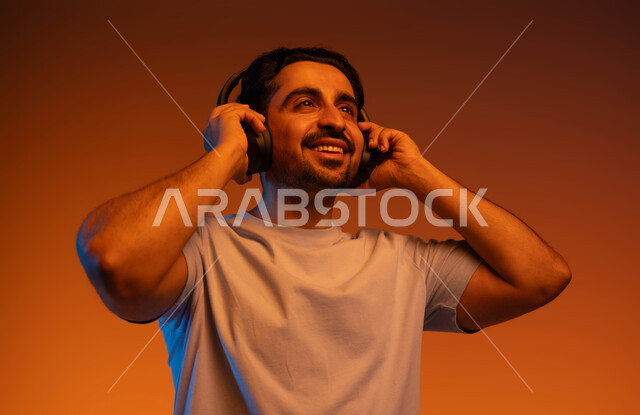 ايماءات وتعابير السرور والطرب، قضاء أوقات ممتعة بسماع الاغاني والمقاطع الصوتية، بورتريه مقرب لشاب عربي خليجي سعودي مبتسم يرتدي الزي الكاجوال يضع سماعة الرأس الصوتية، مفهوم الإصغاء والاستماع بإستخدام تقنية الهيدفون، خلفية ملونة