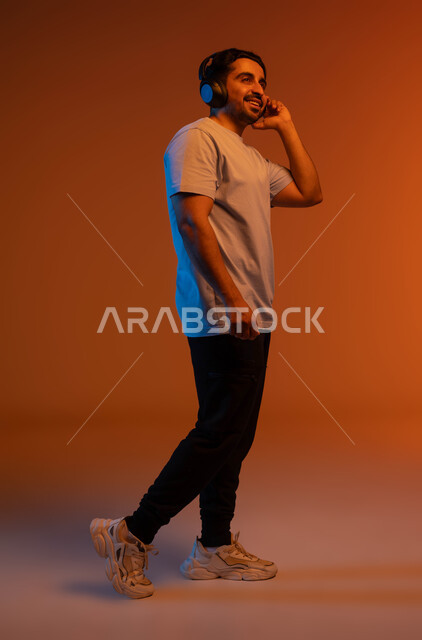 تعابير وايماءات التجول والذهاب الى مكان ما، بورتريه لشاب عربي خليجي سعودي مبتسم يرتدي الزي الكاجوال يضع سماعة الرأس الصوتية، مفهوم الإصغاء والاستماع بإستخدام تقنية الهيدفون، الاستمتاع بسماع الاغاني والمقاطع الصوتية، صورة الجسم بالكامل، خلفية ملونة