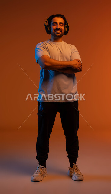 النظر الى الكاميرا والوقوف باستقامة وايدي متقاطعة، بورتريه لشاب عربي خليجي سعودي مبتسم يرتدي الزي الكاجوال يضع سماعة الرأس الصوتية، مفهوم الإصغاء والاستماع بإستخدام تقنية الهيدفون، الاستمتاع بسماع الاغاني والمقاطع الصوتية، صورة الجسم بالكامل، خلفية ملونة