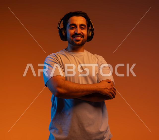 قضاء أوقات ممتعة بسماع الاغاني والمقاطع الصوتية، الوقوف باستقامة وايدي متقاطعة والنظر الى الكاميرا، بورتريه مقرب لشاب عربي خليجي سعودي مبتسم يرتدي الزي الكاجوال يضع سماعة الرأس الصوتية، مفهوم الإصغاء والاستماع بإستخدام تقنية الهيدفون، خلفية ملونة