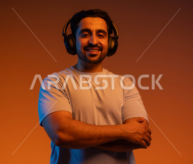الوقوف باستقامة وايدي متقاطعة والنظر الى الكاميرا، قضاء أوقات ممتعة بسماع الاغاني والمقاطع الصوتية، بورتريه مقرب لشاب عربي خليجي سعودي مبتسم يرتدي الزي الكاجوال يضع سماعة الرأس الصوتية، مفهوم الإصغاء والاستماع بإستخدام تقنية الهيدفون، خلفية ملونة