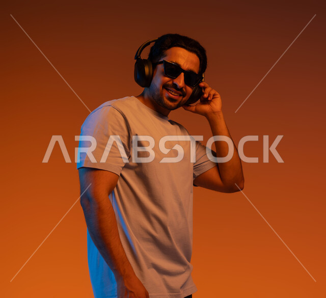 تعابير وايماءات السرور لسماع الأغنية، بورتريه مقرب لشاب عربي خليجي سعودي يرتدي الزي الكاجوال والنظارات الشمسية يضع سماعة رأس في استديو صوتي, الإستماع للأغاني بإستخدام الهيدفون، مواهب غنائية فنية, خلفية ملونة