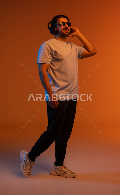 الاندماج والتفاعل مع الانغام الموسيقية، تعابير وايماءات السرور لسماع الأغنية، بورتريه لشاب عربي خليجي سعودي يرتدي الزي الكاجوال والنظارات الشمسية يضع سماعة رأس في استديو صوتي, الإستماع للأغاني بإستخدام الهيدفون، مواهب غنائية فنية, صورة لكامل الجسم، خلفية ملونة