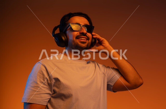 إيماءات وتعابير السرور لسماع الأغنية، بورتريه مقرب لشاب عربي خليجي سعودي يرتدي الزي الكاجوال والنظارات الشمسية يضع سماعة رأس في استديو صوتي, الإستماع للأغاني بإستخدام الهيدفون، مواهب غنائية فنية, خلفية ملونة