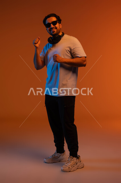 الإستماع للأغاني بإستخدام الهيدفون، الاندماج والتفاعل مع الانغام الموسيقية، تعابير وايماءات السرور لسماع الأغنية، بورتريه لشاب عربي خليجي سعودي يرتدي الزي الكاجوال والنظارات الشمسية يضع سماعة رأس في استديو صوتي, مواهب غنائية فنية, صورة لكامل الجسم، خلفية ملونة