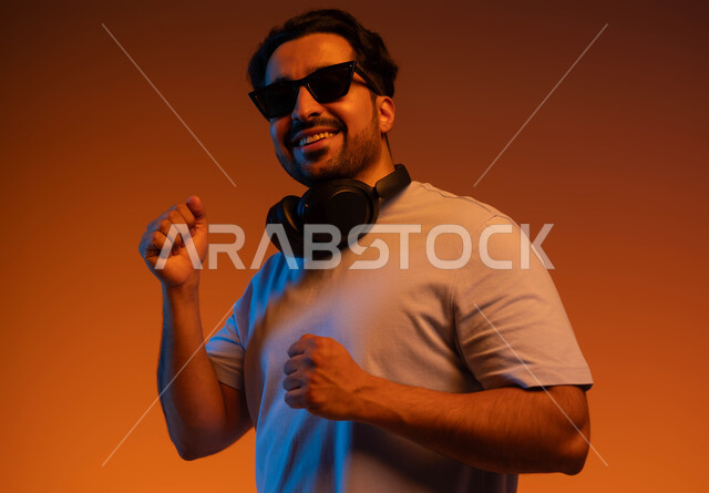 مواهب غنائية فنية, إيماءات وتعابير الاندماج مع الأغنية، بورتريه مقرب لشاب عربي خليجي سعودي يرتدي الزي الكاجوال والنظارات الشمسية يضع سماعة رأس في استديو صوتي, الإستماع للأغاني بإستخدام الهيدفون، خلفية ملونة