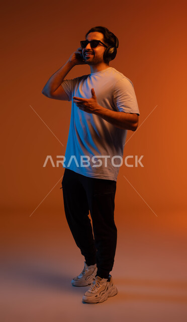 التفاعل والاندماج مع الانغام الموسيقية، تعابير وايماءات السرور لسماع الأغنية، بورتريه لشاب عربي خليجي سعودي يرتدي الزي الكاجوال والنظارات الشمسية يضع سماعة رأس في استديو صوتي, الإستماع للأغاني بإستخدام الهيدفون، مواهب غنائية فنية, صورة لكامل الجسم، خلفية ملونة
