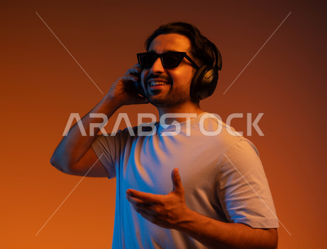 إيماءات وتعابير الاندماج مع الأغنية، بورتريه مقرب لشاب عربي خليجي سعودي يرتدي الزي الكاجوال والنظارات الشمسية يضع سماعة رأس في استديو صوتي, الإستماع للأغاني بإستخدام الهيدفون، مواهب غنائية فنية, خلفية ملونة