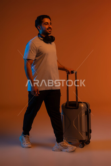 تعابير المشي و الاستعداد للانطلاق وبدء الرحلة، الاستمتاع في قضاء إجازة الصيف، بورتريه لشاب عربي خليجي سعودي مبتسم يرتدي الزي الكاجوال يمسك بيده حقيبة السفر, الاستماع للأغاني في الجولات سياحية باستخدام الهيدفون، صورة الجسم بالطول الكامل ، خلفية ملونة
