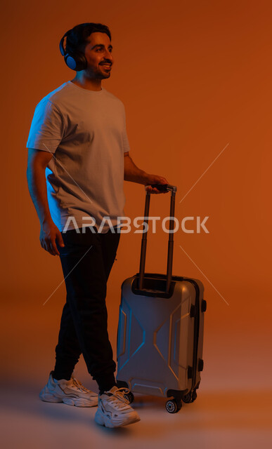 ايماءات المشي و الاستعداد للانطلاق وبدء الرحلة، الاستمتاع في قضاء إجازة الصيف، بورتريه لشاب عربي خليجي سعودي مبتسم يرتدي الزي الكاجوال يمسك بيده حقيبة السفر, الاستماع للأغاني في الجولات سياحية باستخدام الهيدفون، صورة الجسم بالطول الكامل ، خلفية ملونة