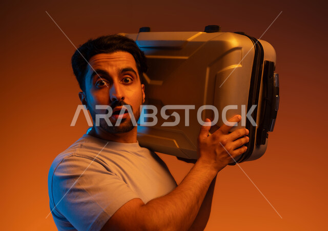 النظر إلى الكاميرا بإيماءات الذهول والدهشة، الذهاب في جولات سياحية ترفيهية في إجازة الصيف, بورتريه مقرب لشاب عربي سعودي خليجي يرتدي الزي الكاجوال يحمل حقيبة السفر على كتفه, الاستعداد لقضاء رحلة صيفية ممتعة, خلفية ملونة