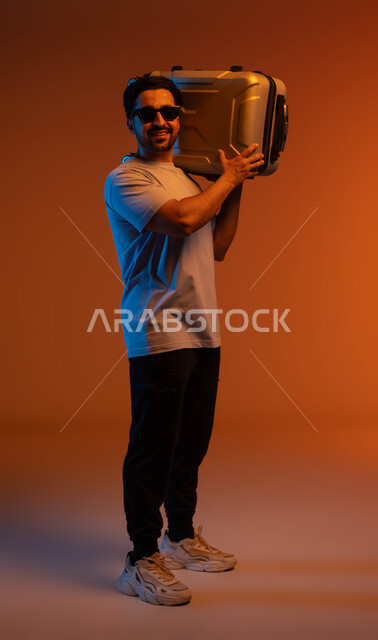 التجهز لقضاء رحلة صيفية ممتعة, نشاطات وفعاليات سياحية، النظر إلى الكاميرا بتعابير الفرح والسرور، الذهاب في جولات ترفيهية في إجازة الصيف, بورتريه لشاب عربي سعودي خليجي يرتدي الزي الكاجوال والنظارات الشمسية يحمل حقيبة السفر على كتفه, صورة لكامل الجسم، خلفية ملونة