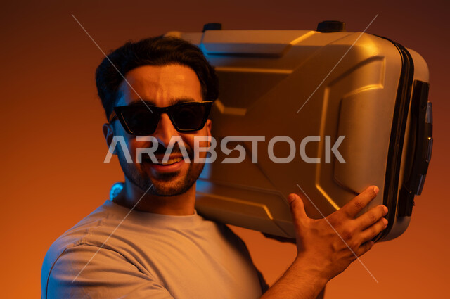 انشطة وفعاليات سياحية، النظر إلى الكاميرا بتعابير الفرح والسرور، الذهاب في جولات ترفيهية في إجازة الصيف, بورتريه مقرب لشاب عربي سعودي خليجي يرتدي الزي الكاجوال والنظارات الشمسية يحمل حقيبة السفر على كتفه, الاستعداد لقضاء رحلة صيفية ممتعة, خلفية ملونة
