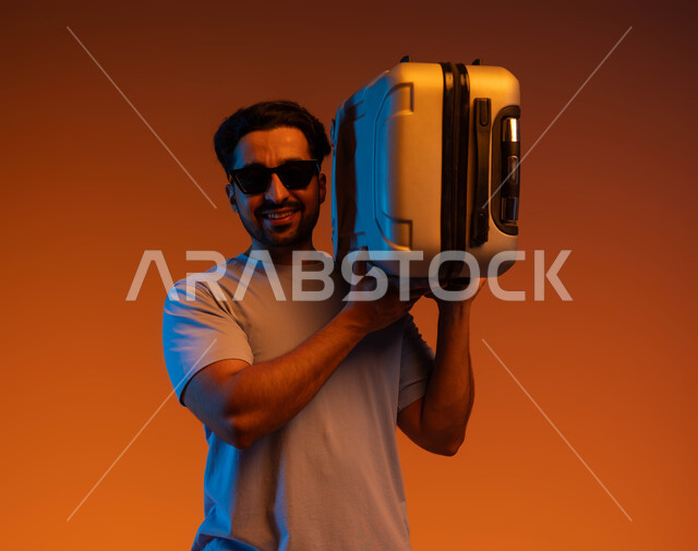 The concept of elegance and attention to external appearance, preparing for an enjoyable summer trip, tourist activities and events, expressions of joy and happiness, going on recreational tours during the summer vacation, a close-up portrait of a young Saudi Arabian Gulf man wearing casual clothes and sunglasses carrying a travel bag on his shoulder, colored background.