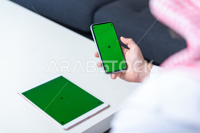 رجل أعمال سعودي خليجي يستخدام هاتفه المحمول بخلفية خضراء كروما ، تقنية و حاسب ، استخدام تقنية السمارت على الهاتف المحمول ليتحكم بالتلفاز