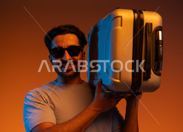The concept of elegance and attention to external appearance, preparing for an enjoyable summer trip, tourist activities and events, expressions of joy and happiness, going on recreational tours during the summer vacation, a close-up portrait of a young Saudi Arabian Gulf man wearing casual clothes and sunglasses carrying a travel bag on his shoulder, colored background.