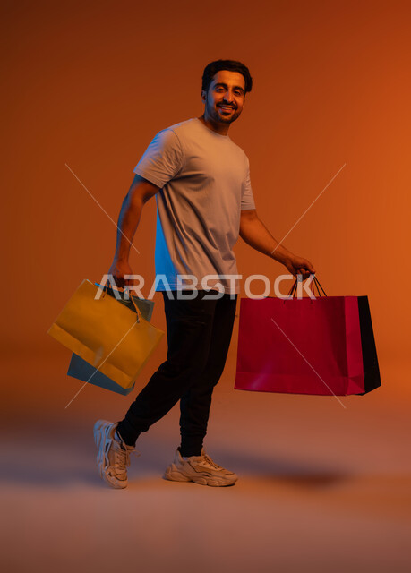 الاستفادة من العروضات والتخفيضات، بورتريه لرجل عربي خليجي سعودي مبتسم يرتدي الزي الكاجوال يحمل بيده أكياس ورقية ملونة بإيماءات السرور والسعادة، المتعة في التسوق واقتناء المستلزمات والهدايا، صورة الجسم بالطول الكامل، خلفية ملونة