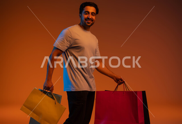المتعة في التسوق واقتناء المستلزمات والهدايا، الاستفادة من العروضات والتخفيضات، بورتريه لرجل عربي خليجي سعودي مبتسم يرتدي الزي الكاجوال يحمل بيده أكياس ورقية ملونة بإيماءات السرور والسعادة، خلفية ملونة