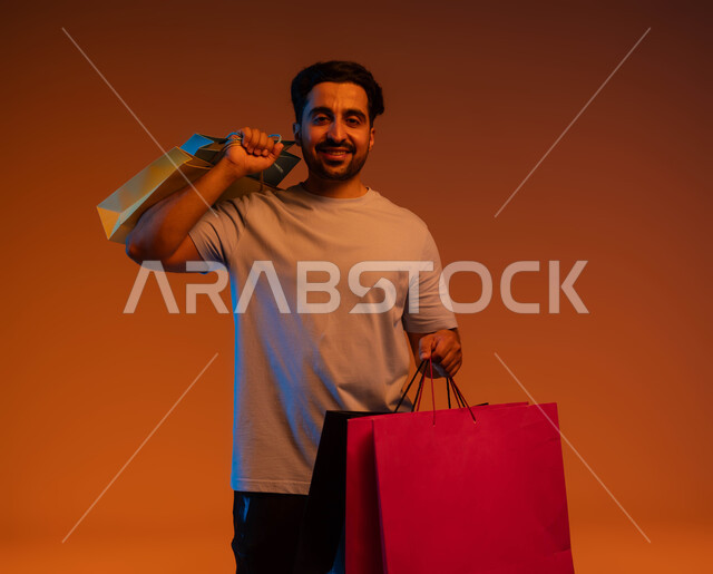 اقتناء المستلزمات والهدايا، تعابير الحماس للتسوق، الاستفادة من عروض وتخفيضات الجمعة البيضاء، بورتريه لرجل عربي خليجي سعودي مبتسم يحمل بيده أكياس ورقية ملونة ينظر إلى الكاميرا بإيماءات السرور والسعادة، خلفية ملونة.