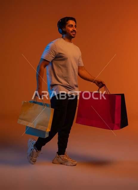 الحسومات و التنزيلات الموسمية، العودة من التسوق بإيماءات السعادة والفرح، الاستفادة من العروض و الخصومات، بورتريه لشاب عربي خليجي سعودي يرتدي سماعة الرأس يحمل بيديه حقائب التسوق، صورة الجسم بالطول الكامل، خلفية ملونة.