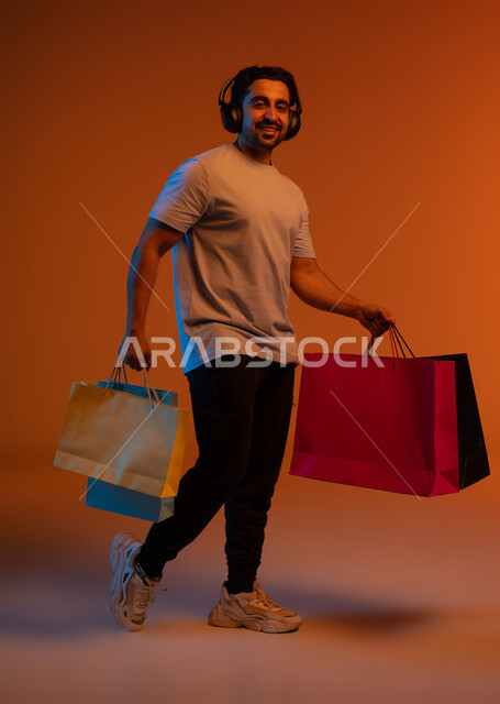 العودة من التسوق بإيماءات السعادة والفرح، التنزيلات الموسمية والحسومات، الاستفادة من العروض و الخصومات، بورتريه لشاب عربي خليجي سعودي ينظر إلى الكاميرا يرتدي سماعة الرأس يحمل بيديه حقائب التسوق، صورة الجسم بالطول الكامل، خلفية ملونة.