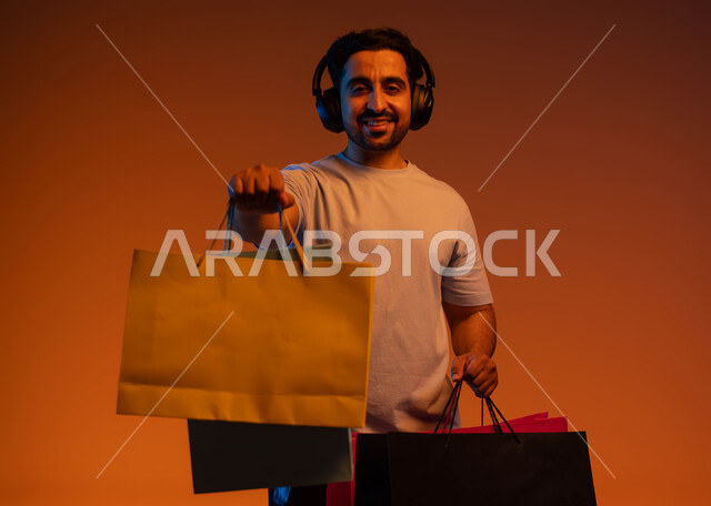 الشعور بالفرح والسعادة، التنزيلات الموسمية والحسومات، الاستفادة من العروض و الخصومات، بورتريه مقرب لشاب عربي خليجي سعودي ينظر إلى الكاميرا يرتدي سماعة الرأس يحمل بيديه حقائب التسوق، الاستمتاع بالشراء و التبضع وسماع الموسيقا، خلفية ملونة.
