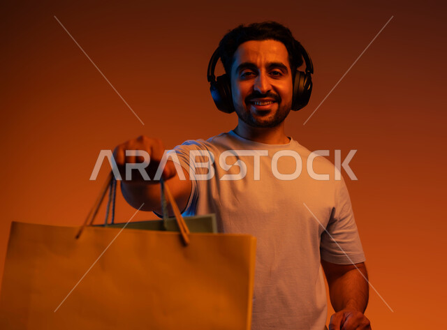 التنزيلات الموسمية والحسومات، الاستفادة من العروض و الخصومات، بورتريه مقرب لشاب عربي خليجي سعودي ينظر إلى الكاميرا يرتدي سماعة الرأس يحمل بيديه حقائب التسوق يقوم برفعها للاعلى، الاستمتاع بالشراء و التبضع وسماع الموسيقا، خلفية ملونة.