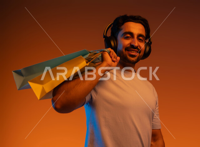 الاستفادة من العروض و الخصومات، بورتريه مقرب لشاب عربي خليجي سعودي ينظر إلى الكاميرا يرتدي سماعة الرأس يحمل بيديه حقائب التسوق، الاستمتاع بالشراء و التبضع وسماع الموسيقا، الحسومات و التنزيلات الموسمية، خلفية ملونة.