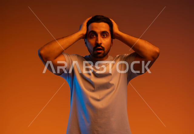 النظر إلى الكاميرا بإيماءات التعجب والدهشة، التفاجؤ والاستغراب من شيء ما، بورتريه مقرب لشاب عربي خليجي سعودي يرتدي الزي الكاجوال و يرفع يديه على رأسه يشعر بالصدمة والذهول والانبهار، تعابير النجاح وتحقيق الأهداف, خلفية ملونة.