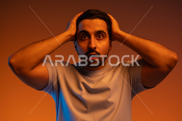 النظر إلى الكاميرا بإيماءات التعجب والدهشة، التفاجؤ والاستغراب من شيء ما، بورتريه مقرب لشاب عربي خليجي سعودي يرتدي الزي الكاجوال و يرفع يديه على رأسه يشعر بالصدمة والذهول والانبهار، تعابير النجاح وتحقيق الأهداف, خلفية ملونة.