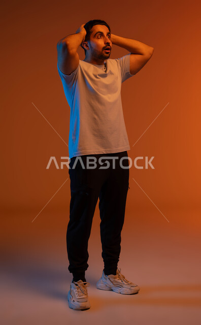 Surprise and astonishment at something, a feeling of shock, astonishment and astonishment, portrait of a Saudi Gulf Arab young man wearing casual clothes and raising his hands to his head with gestures of wonder and astonishment, expressions of success and achieving goals, full-length body portrait, colored background